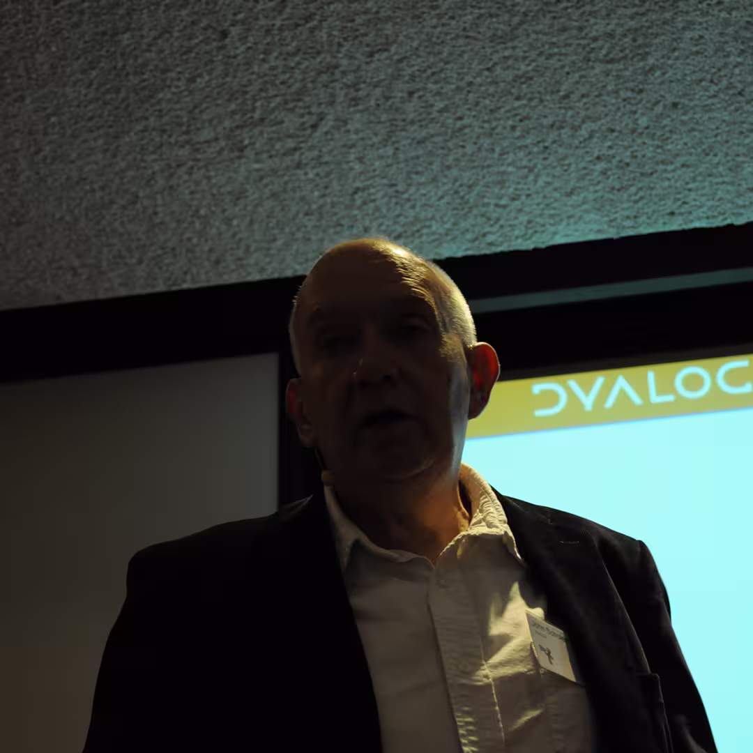 John Scholes speaking at the Dyalog '14 event in Eastbourne, UK. He is standing in front of a presentation screen displaying the Dyalog logo, wearing a white shirt and dark jacket.