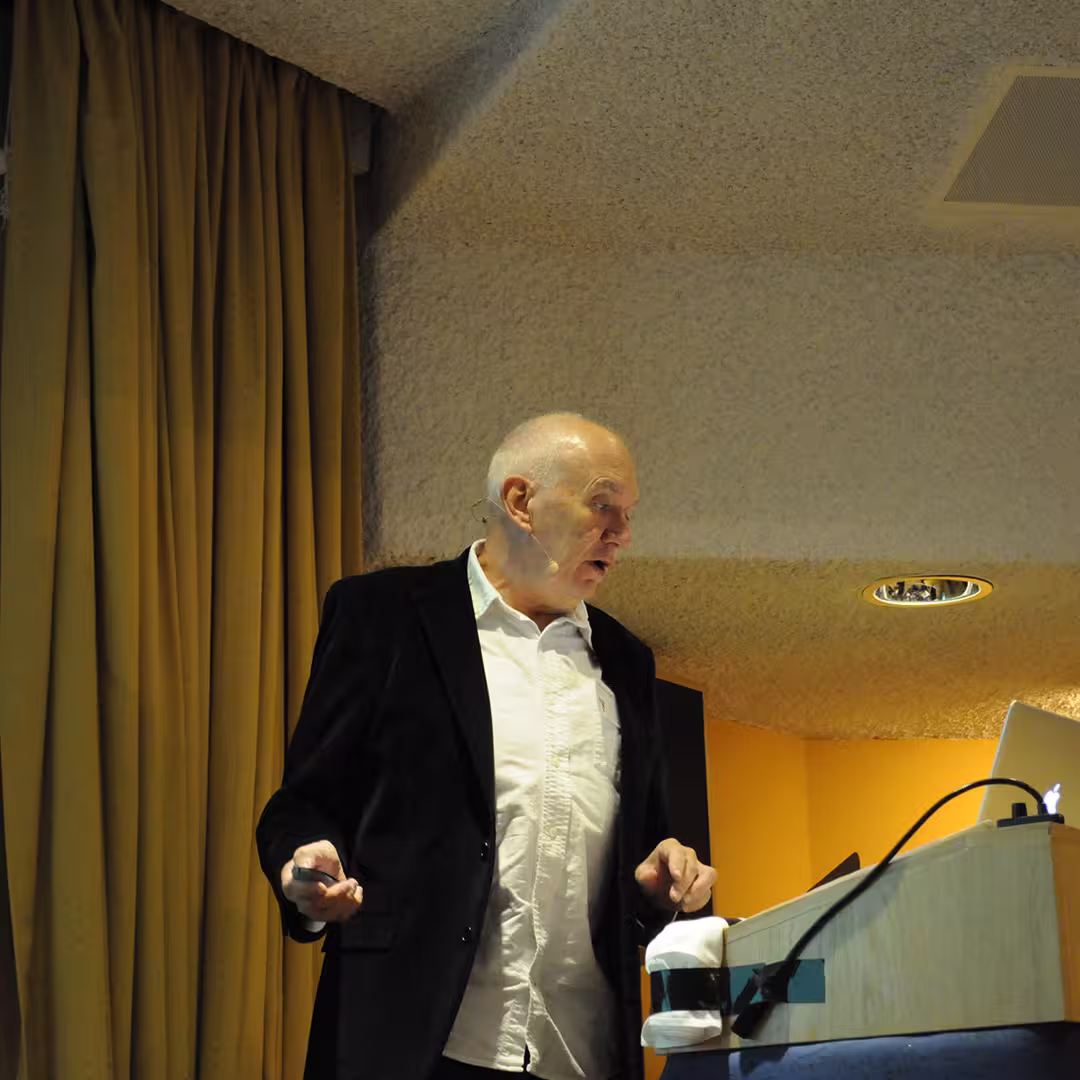 John Scholes presenting at the Dyalog '14 event in Eastbourne, UK. He stands at a lectern with a microphone headset, gesturing while speaking, with a laptop and a yellow curtain in the background.