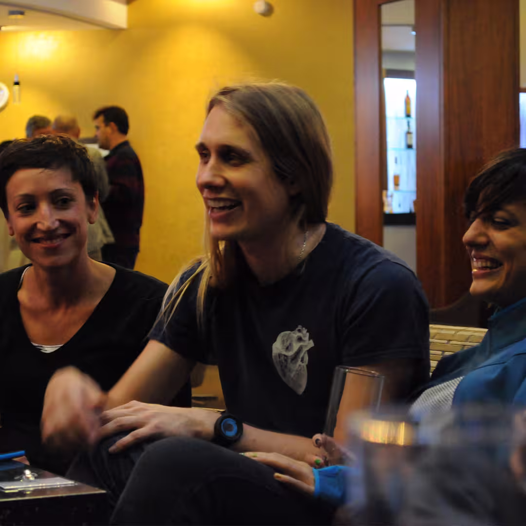 Three Dyalog '14 delegates seated together in a lounge area, smiling and engaged in conversation. The background features other attendees and a bar area.