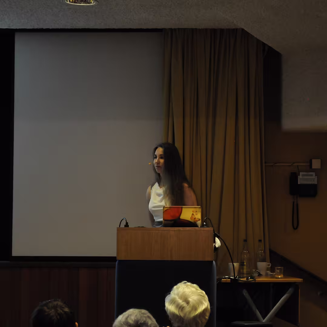 A Dyalog '14 delegate presenting at a lectern in Eastbourne, UK. The speaker, a young woman with long dark hair, is wearing a white top and using a laptop with a colorful cover. The background features a yellow curtain and a blank projection screen.