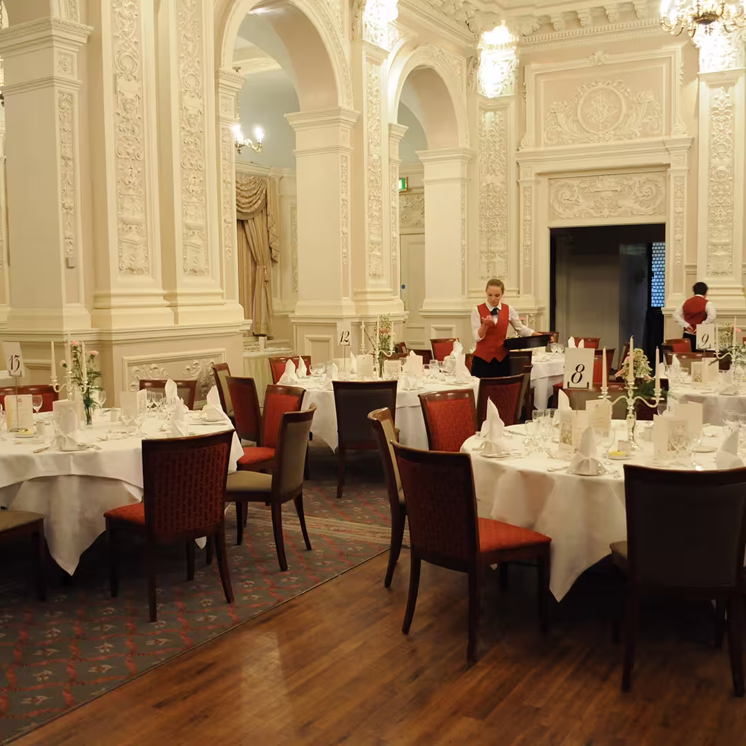 Elegant dining room set up for the Dyalog '14 event in Eastbourne, UK. The room features ornate white plasterwork, chandeliers, and round tables with white tablecloths, floral centerpieces, and neatly folded napkins. Staff in red waistcoats are preparing the tables.
