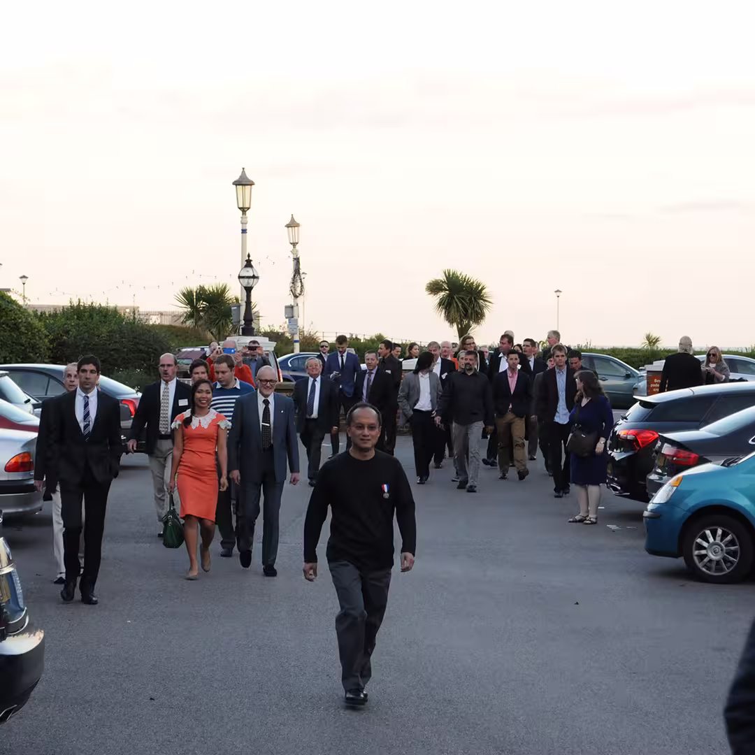 Roger Hui leading a group of Dyalog '14 delegates walking along a street in Eastbourne, UK, dressed in formal and semi-formal attire. Cars are parked on either side, with street lamps and palm trees in the background.