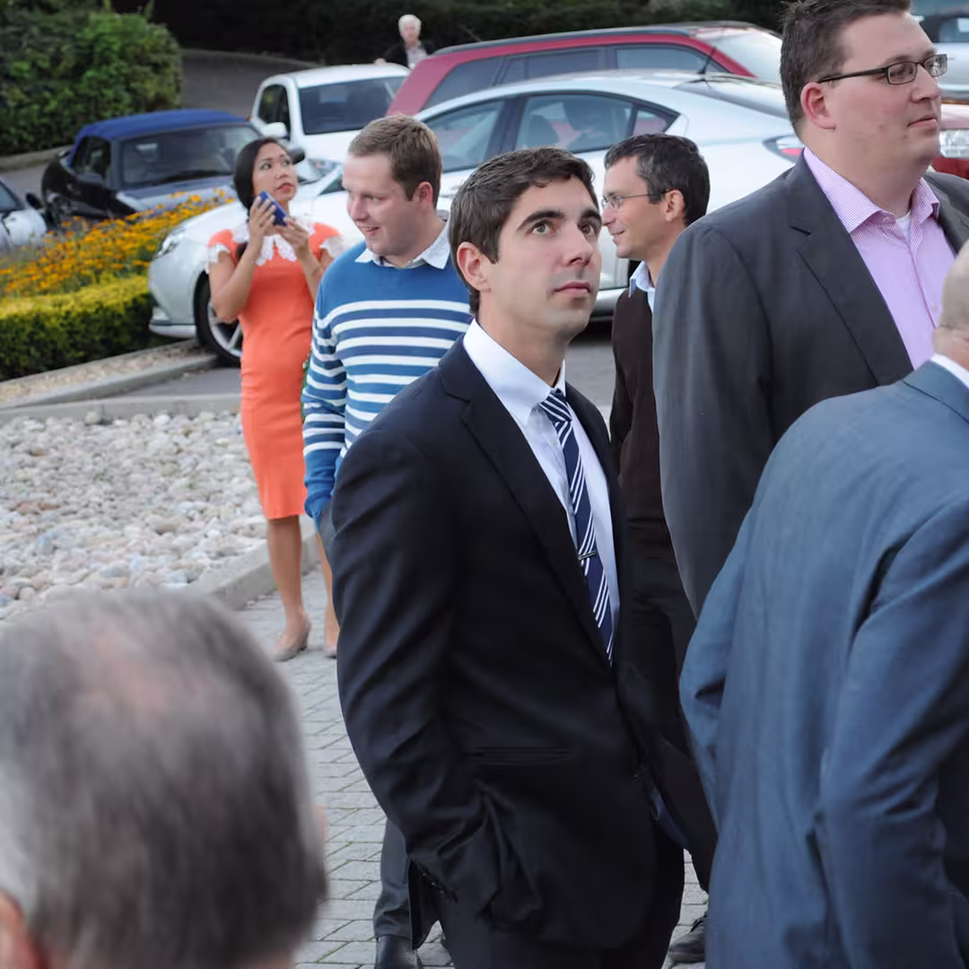 Dyalog '14 delegates gathered outside in Eastbourne, UK. A man in a dark suit and striped tie stands in the center, looking up, while others around him engage in conversation. Cars and landscaping are visible in the background.
