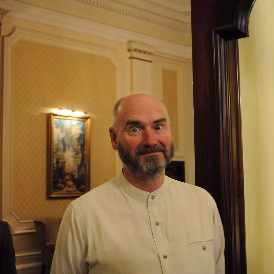 Stephen Taylor at the Dyalog '14 event in Eastbourne, UK, looking at the camera with a light-colored, buttoned shirt. He has a beard and a playful expression, standing in a traditionally decorated room with a framed painting in the background.
