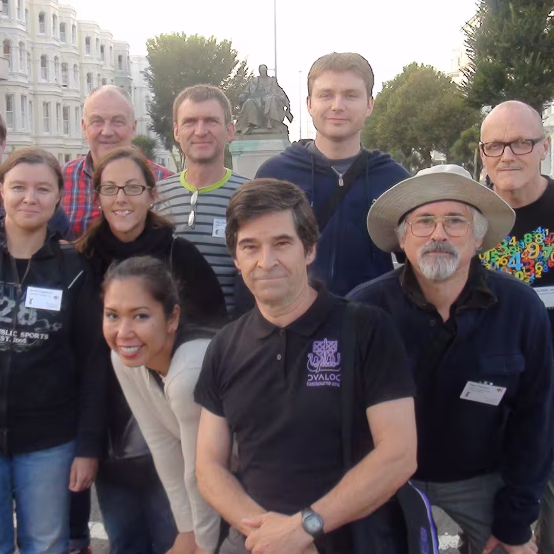 Group of Dyalog '14 attendees posing outdoors in Eastbourne, UK. Dan Baronet is in the middle, wearing a black Dyalog shirt, and Ray Cannon is on his left, wearing a wide-brimmed hat and glasses. The background features a statue and tree-lined street.