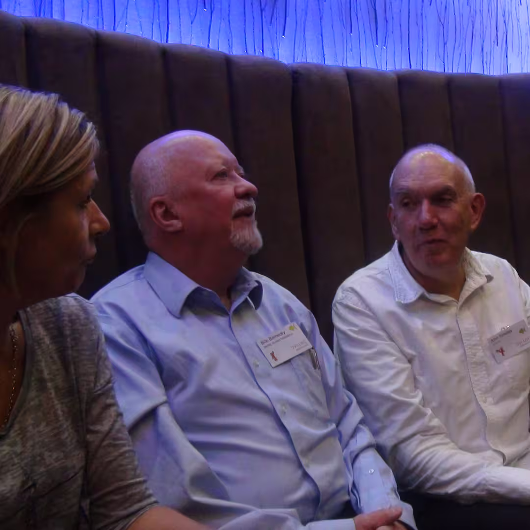 John Scholes (right) seated with two other Dyalog '14 delegates in Eastbourne, UK. They are engaged in conversation, seated in front of a booth with a textured blue-lit wall.