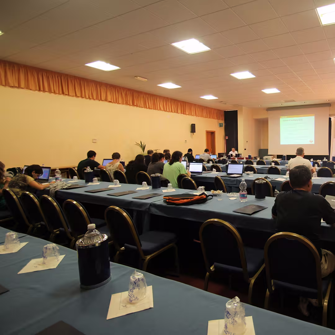 A conference room at the Dyalog '15 event in Sicily, Italy, showing delegates seated at long tables covered with blue tablecloths. Each table has water bottles, glasses, and notepads. Attendees are working on laptops while facing the front of the room, where a presentation slide titled "Research Summary (Q2)" is projected onto a screen. The room has a yellowish wall with a long orange curtain near the ceiling, and the ceiling is fitted with bright fluorescent lights.