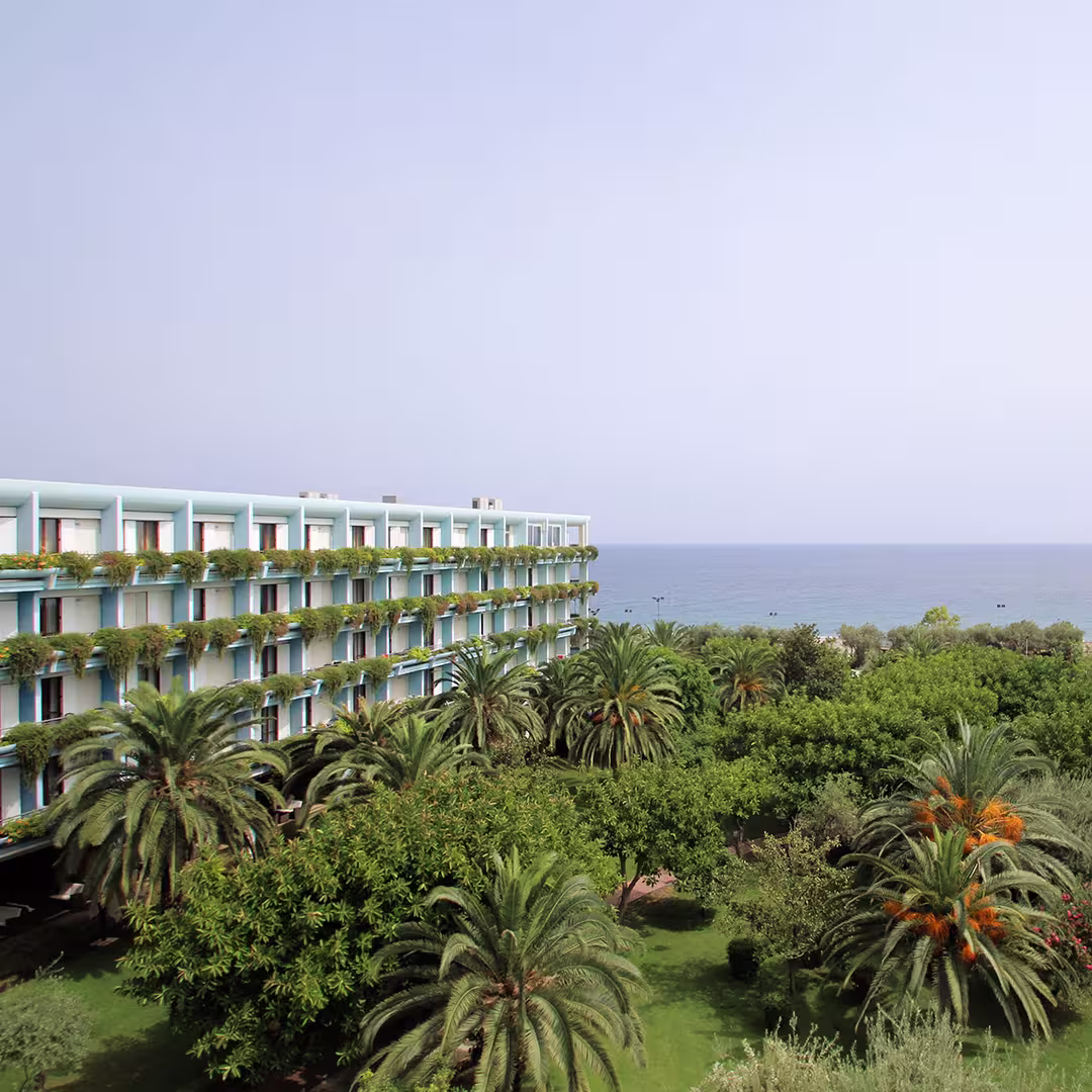 A view of the Dyalog '15 event venue in Sicily, Italy, showing a modern, multi-story hotel with white walls and blue balconies. Each balcony has lush green plants cascading over the railings. In the foreground, there is a vibrant garden filled with tall palm trees and other green foliage. In the background, the calm, blue Mediterranean Sea stretches out to the horizon under a clear sky.