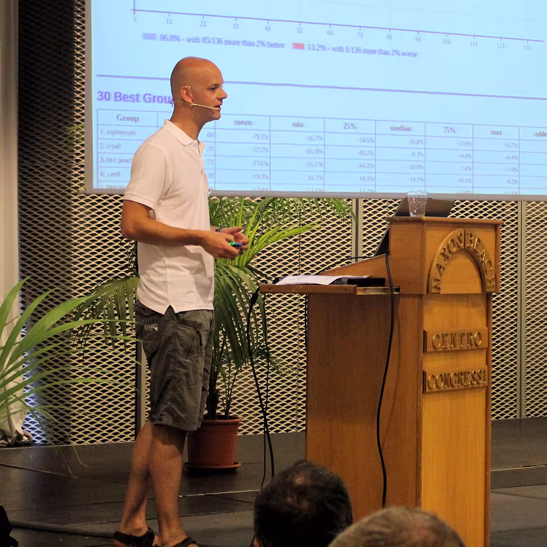 Jay Foad presenting at the Dyalog '15 event in Sicily, Italy. He stands barefoot on stage, wearing a white polo shirt and grey cargo shorts, speaking into a headset microphone while holding a presentation remote. Behind him, a large screen displays a detailed table and chart titled "30 Best Groups." A wooden podium with the words "Marinella Centro Congressi" engraved on it holds a laptop and a glass of water. A potted palm plant is placed beside the stage.