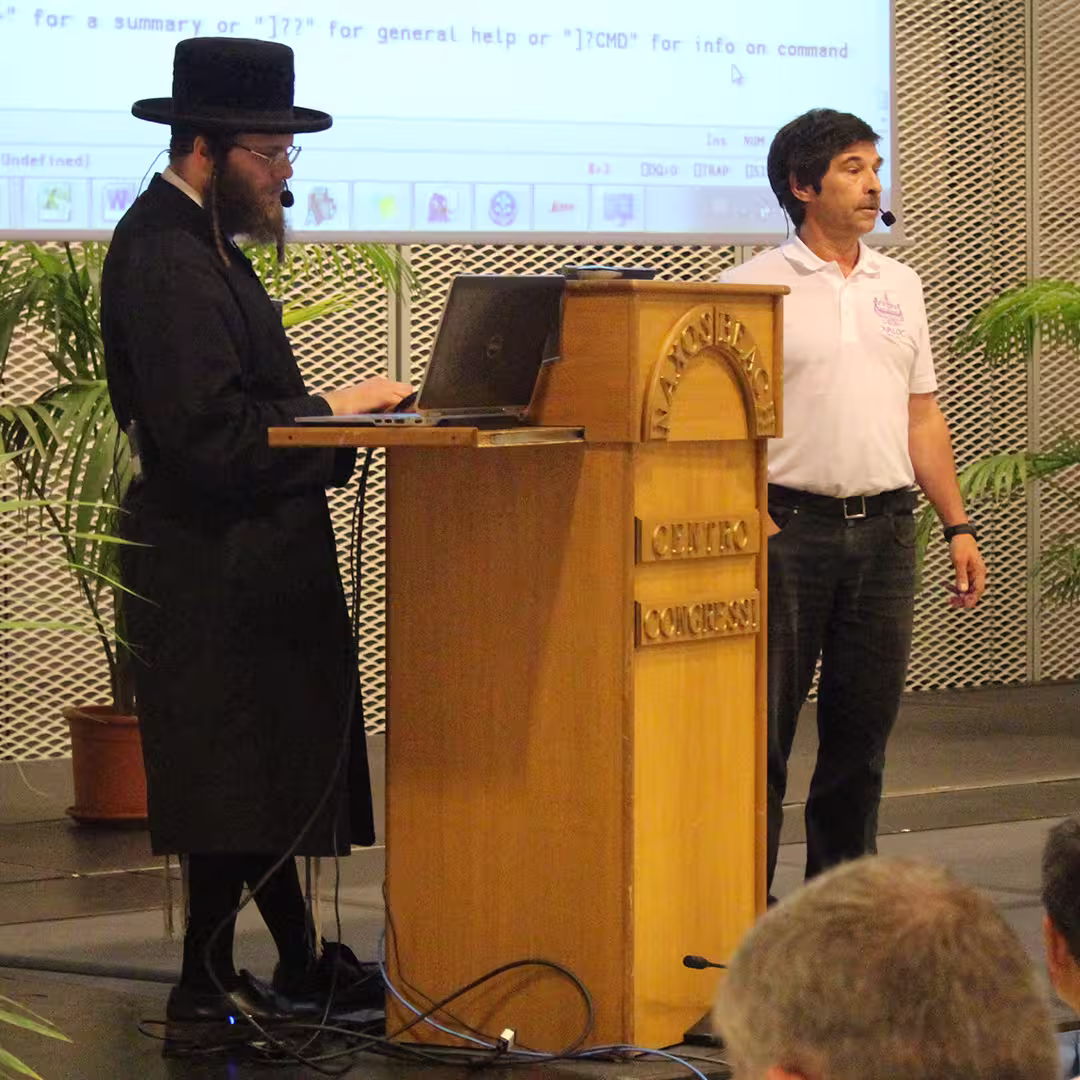 Adám Brudzewsky and Dan Baronet presenting at the Dyalog '15 event in Sicily, Italy. Adám Brudzewsky, dressed in traditional Orthodox Jewish attire, including a black hat and long black coat, is working on a laptop at the wooden podium labeled "Marinella Centro Congressi." Dan Baronet, wearing a white polo shirt and dark trousers, stands to the right, speaking into a headset microphone. The background features a large screen displaying an APL workspace and potted palm plants.