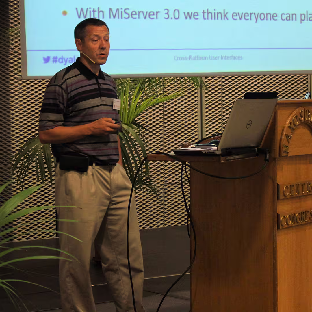 Morten Kromberg presenting at the Dyalog '15 event in Sicily, Italy. He stands beside the wooden podium labeled "Marinella Centro Congressi," wearing a striped purple and black polo shirt and beige trousers. Morten is speaking into a headset microphone while holding a presentation remote. Behind him, a large screen displays a slide about MiServer 3.0 with the hashtag "#dyalog15" and the phrase "Cross-Platform User Interfaces." Potted palm plants decorate the stage area.