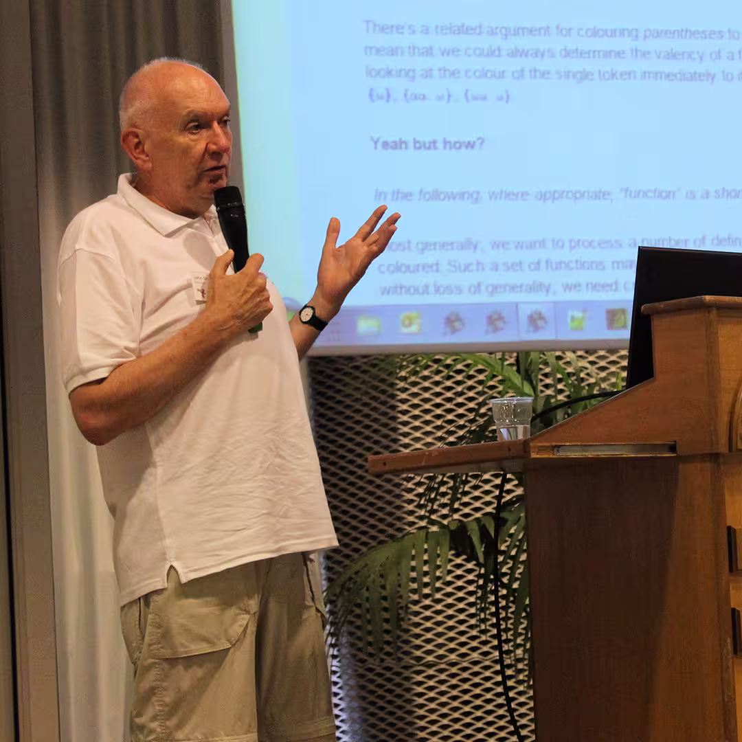 John Scholes presenting at the Dyalog '15 event in Sicily, Italy. He stands beside the wooden podium labeled "Marinella Centro Congressi," wearing a white polo shirt and beige cargo shorts. John is holding a microphone and gesturing with his left hand while speaking. Behind him, a large screen displays a slide discussing the colouring of parentheses in programming, with the heading "Yeah but how?" and several lines of technical text. A potted palm plant is visible near the stage.