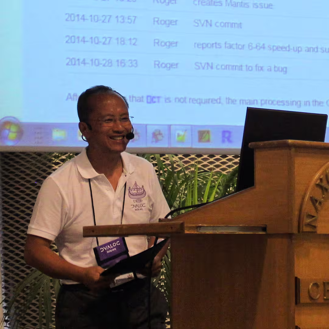 Roger Hui presenting at the Dyalog '15 event in Sicily, Italy. He stands beside the wooden podium labeled "Marinella Centro Congressi," wearing a white Dyalog polo shirt and dark trousers. Roger is smiling while holding a black folder and speaking into a headset microphone. Behind him, a large screen displays a list of SVN commits and bug fixes, with several timestamps and the name "Roger" appearing multiple times. Potted palm plants decorate the stage area.
