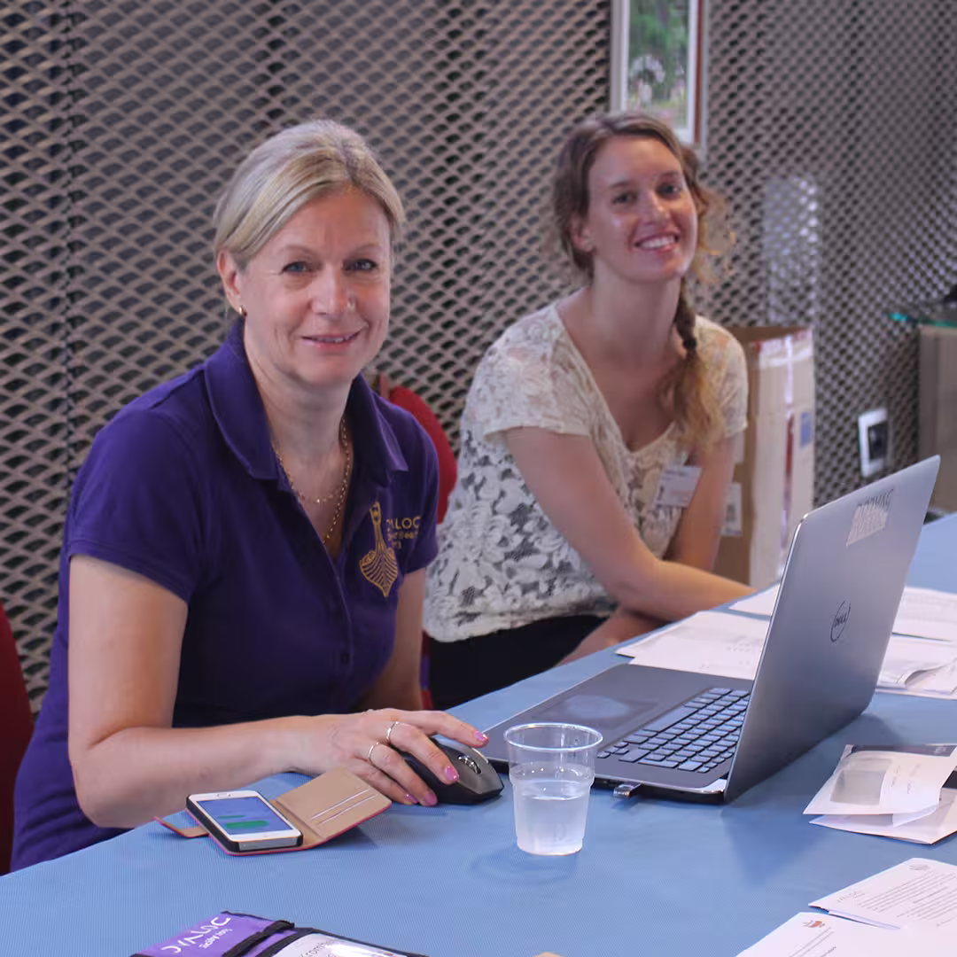 Karen Shaw (left) seated at the registration desk during the Dyalog '15 event in Sicily, Italy. She is wearing a purple Dyalog polo shirt and smiling at the camera while using a computer mouse. A smartphone in a brown case lies on the table beside her. To her right, another Dyalog staff member with long blonde hair tied in a side braid, wearing a white lace top, is also smiling. The desk is covered with a blue tablecloth and has a laptop, a glass of water, papers, and a Dyalog-branded lanyard. The background features a metal mesh wall and a cardboard box.