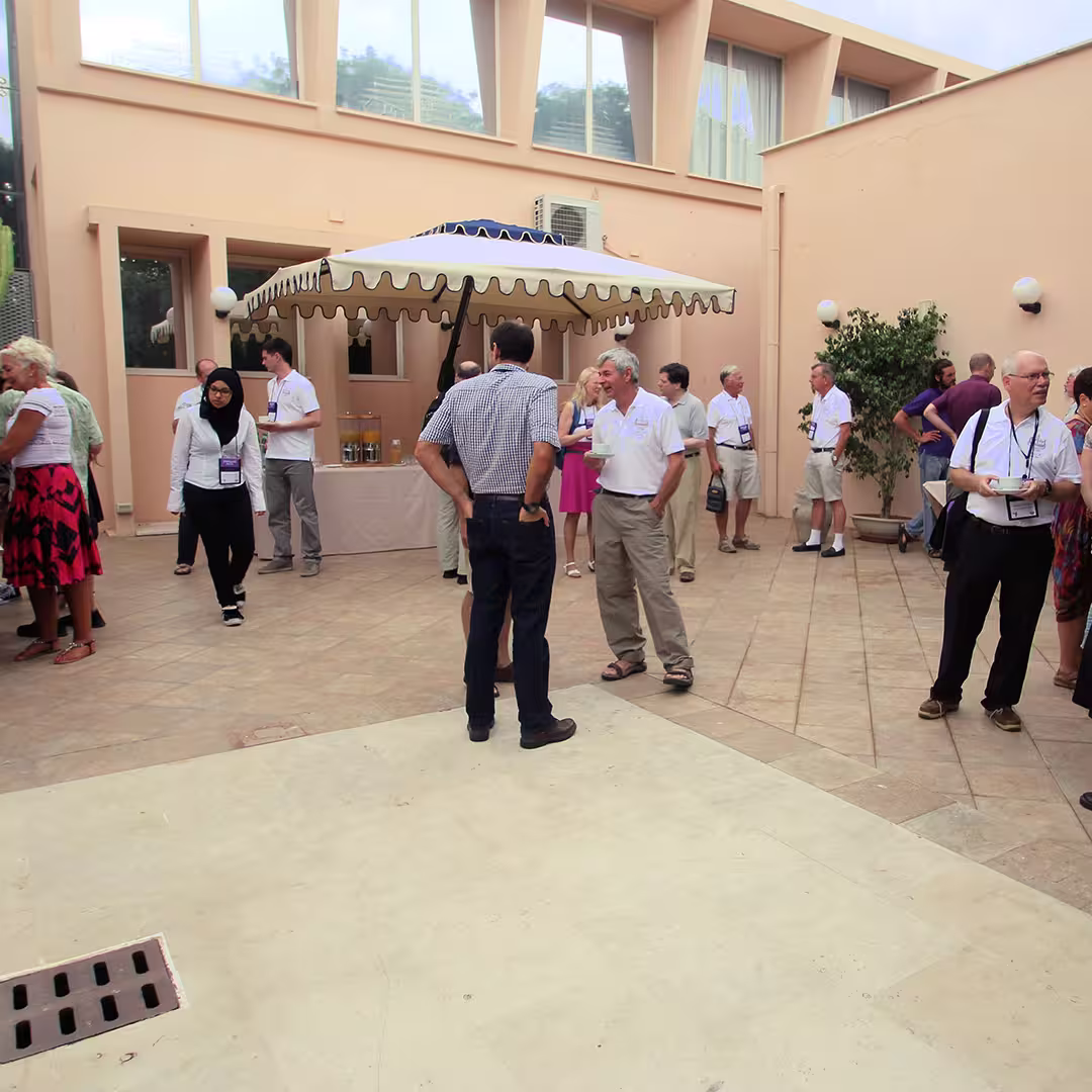 Outdoor networking event during the Dyalog '15 conference in Sicily, Italy. Delegates are mingling in a courtyard with peach-colored walls and large windows. A white canopy with scalloped edges shades a table with refreshments, including bottles of orange juice and water. Attendees are engaged in conversation, some wearing Dyalog-branded lanyards. A woman in a black hijab and white shirt walks across the courtyard, while others stand in small groups chatting. The floor is paved with beige tiles, and a potted plant decorates one corner.