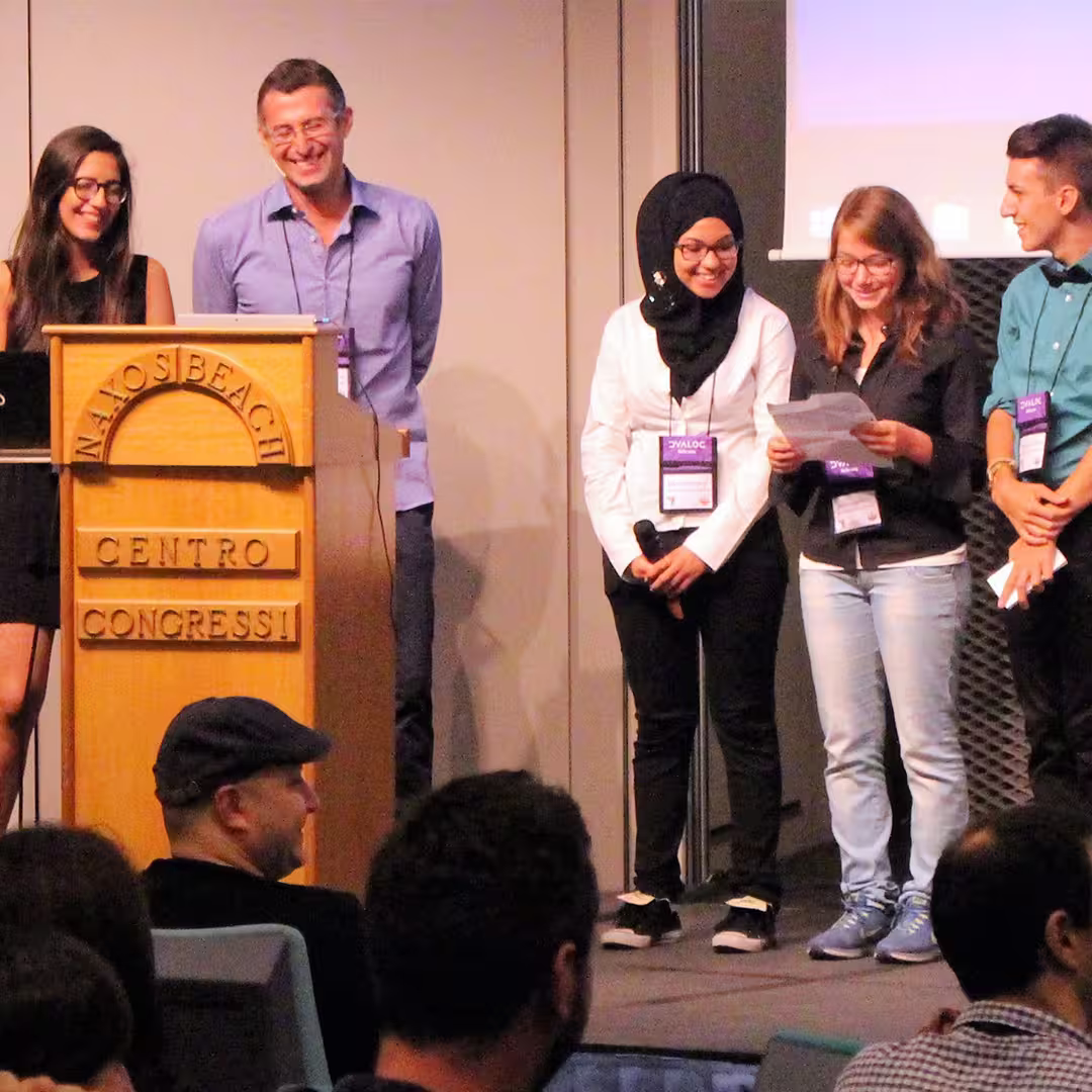 A group of five people on stage during the Dyalog '15 event in Sicily, Italy, standing beside the wooden podium labeled "Naxos Beach Centro Congressi." Two women and one man hold microphones and papers while smiling and presenting. One woman is wearing a black dress, another is in a white shirt with a black hijab, and the third is in a black jacket with light blue jeans. The two men are dressed in a light blue shirt and a turquoise shirt with a black bow tie. Audience members are visible in the foreground, some wearing caps and looking towards the stage.