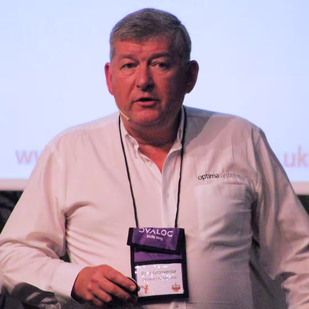Paul Grosvenor of Optima Systems presenting at the Dyalog '15 event in Sicily, Italy. He is wearing a white Optima Systems-branded shirt and a purple Dyalog lanyard with a name badge that reads "Paul Grosvenor, Optima Systems Ltd." Paul is speaking into a headset microphone while standing in front of a projection screen displaying a partially visible web address ending in ".uk."