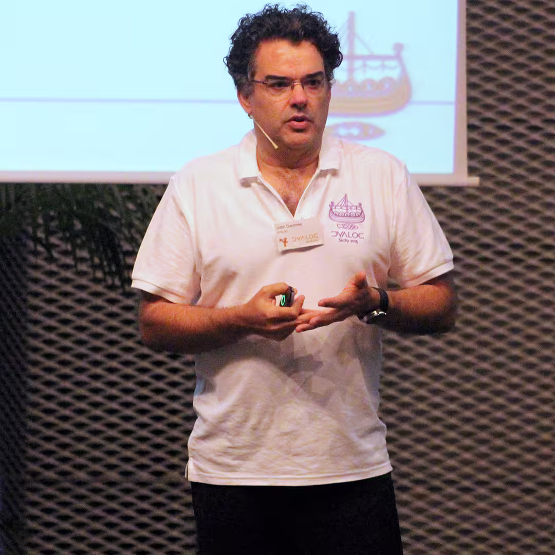 John Daintree presenting at the Dyalog '15 event in Sicily, Italy. He is wearing a white Dyalog-branded polo shirt with the conference logo and a name badge. John is speaking into a headset microphone while holding a presentation remote in his right hand and gesturing with his left. Behind him, a projection screen displays a slide featuring a stylized Viking ship logo. A potted palm plant is visible near the stage.