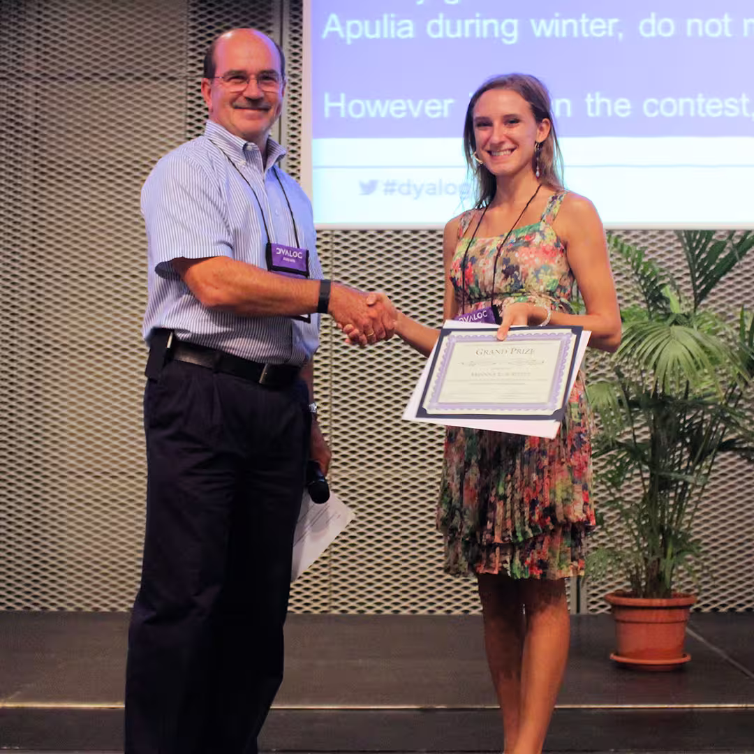 Award presentation at the Dyalog '15 event in Sicily, Italy. A man wearing a light blue striped shirt, dark trousers, and a Dyalog conference lanyard shakes hands with a young woman in a floral dress. She is holding a framed certificate labeled "Grand Prize" while smiling at the camera. The stage background features a projection screen with partially visible text, including the hashtag "#dyalog15," and a potted palm plant stands nearby.