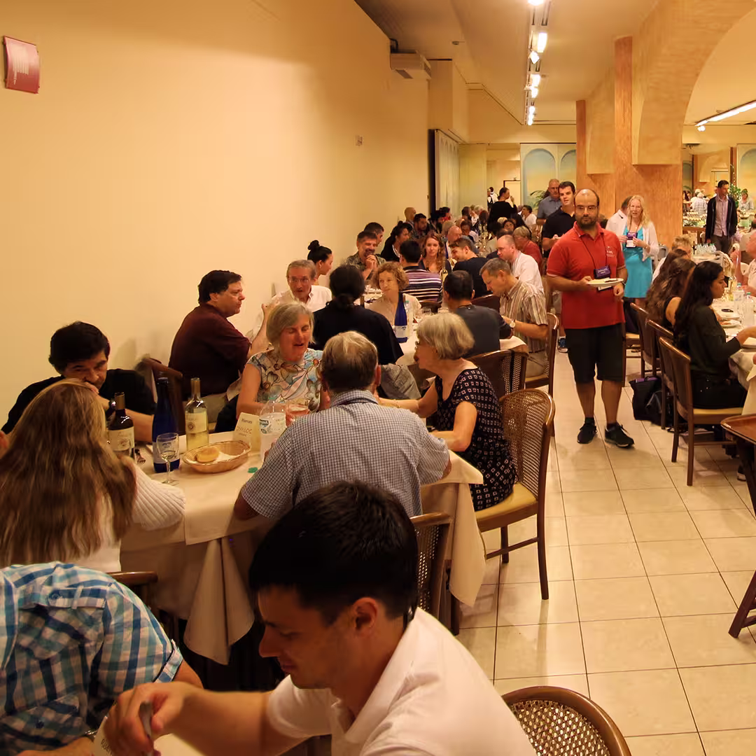 Dinner gathering during the Dyalog '15 event in Sicily, Italy. Delegates are seated at long tables covered with white tablecloths, engaged in conversation while enjoying a meal. Bottles of wine, water, and bread baskets are placed along the tables. In the background, more attendees are seated, while a man in a red shirt with a Dyalog lanyard walks through the room carrying a plate. The dining area has warm yellow walls, arched columns, and soft overhead lighting.