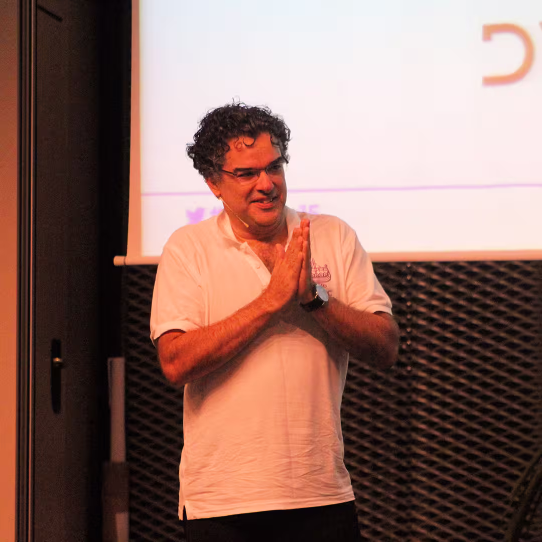 John Daintree on stage at the Dyalog '15 event in Sicily, Italy. He is wearing a white Dyalog-branded polo shirt and glasses, smiling while clasping his hands together. Behind him, a projection screen displays partially visible text and the hashtag "#dyalog15." The background also features a metal mesh wall and a rolled-up poster or banner leaning against the side of the stage.