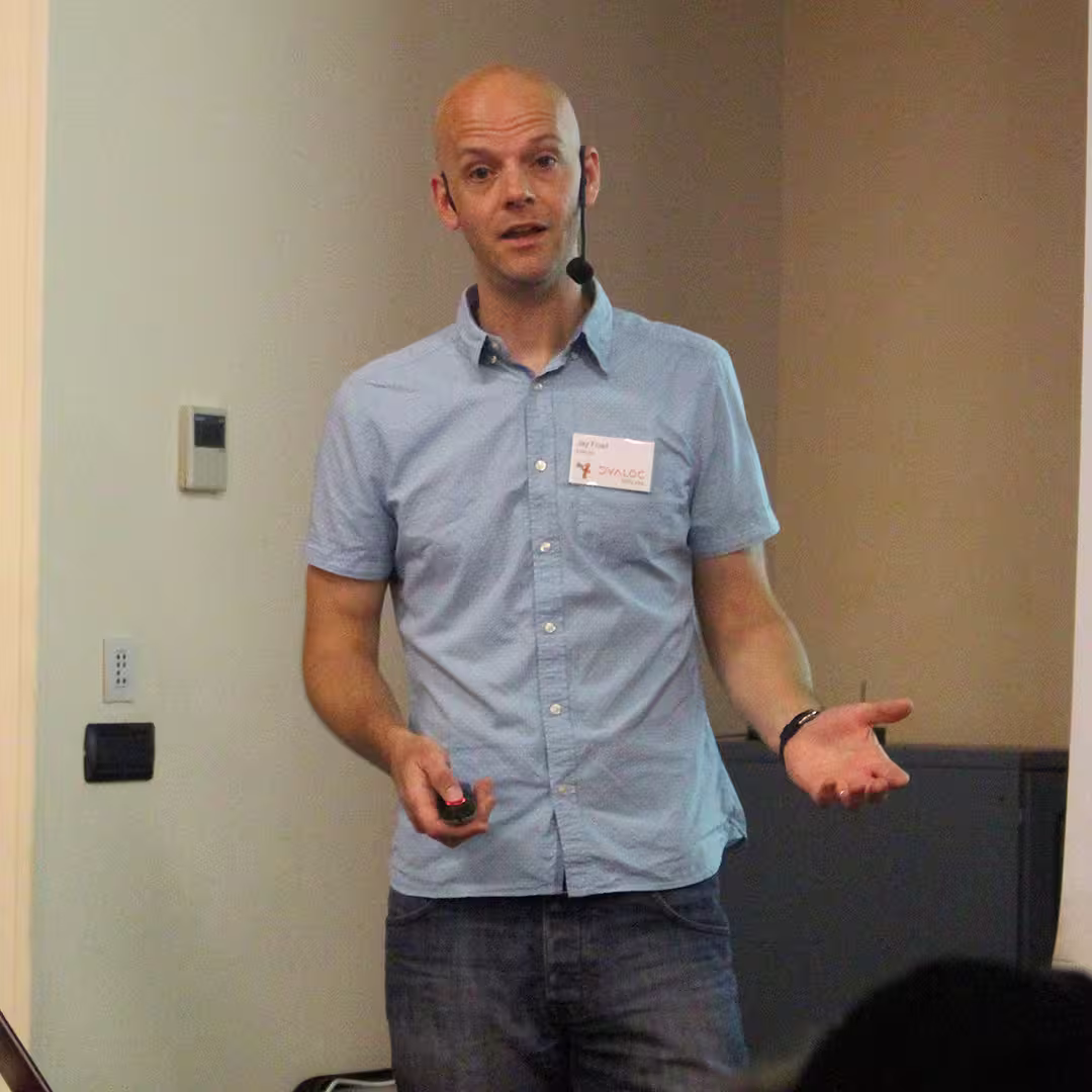 Jay Foad presenting at the Dyalog '15 event in Sicily, Italy. He is wearing a light blue short-sleeved button-up shirt and a Dyalog name badge. Jay is speaking into a headset microphone while holding a presentation remote in his right hand and gesturing with his left. The background features a plain wall with a thermostat and electrical outlet visible.
