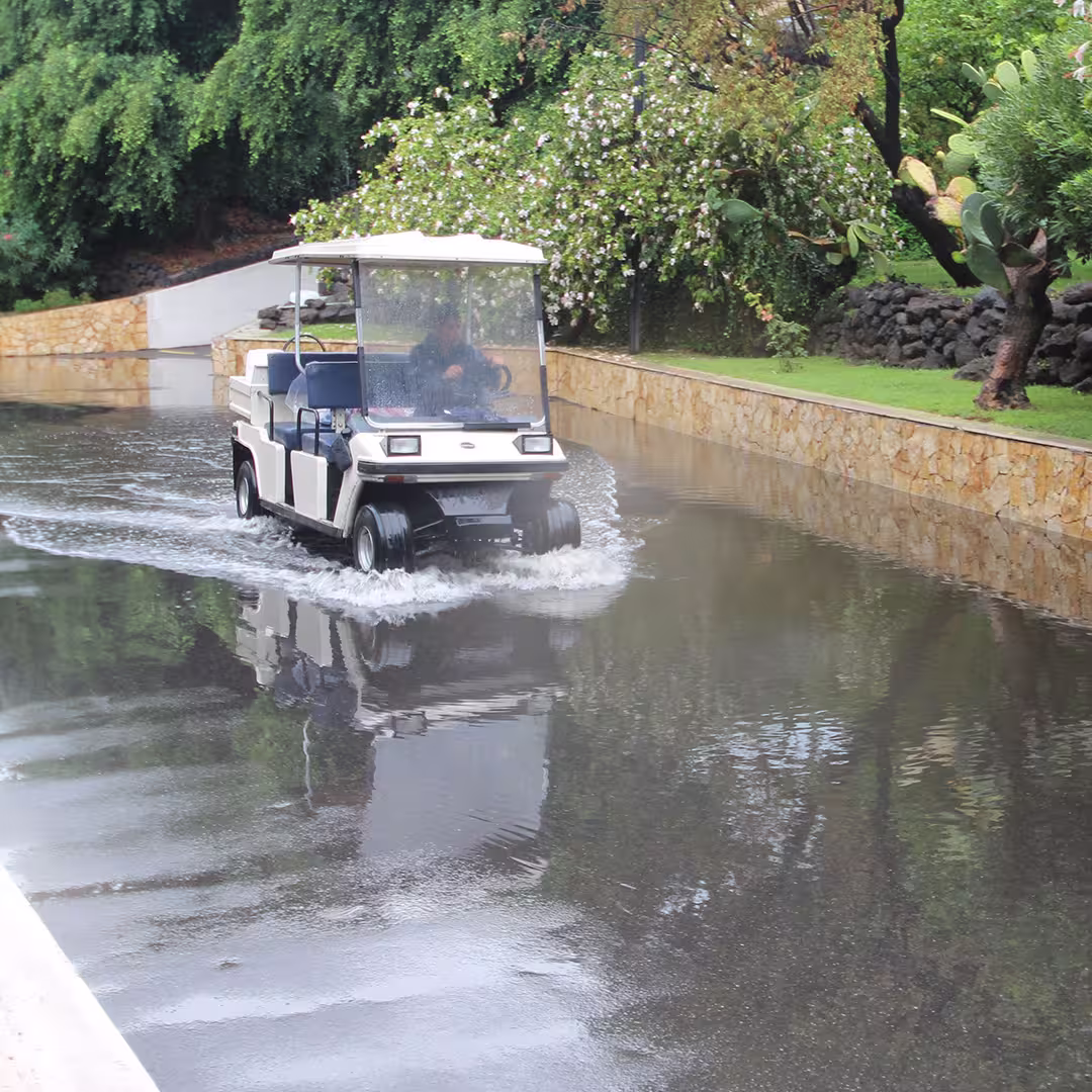 A golf cart driving through a flooded pathway at the Dyalog '15 event venue in Sicily, Italy. The cart is white with a transparent rain cover, and two people are seated inside. Water splashes around the tires as they move forward. The pathway is lined with a stone wall on one side, with lush greenery, flowering bushes, and cactus plants visible along the edge. The sky appears overcast, indicating recent rainfall.