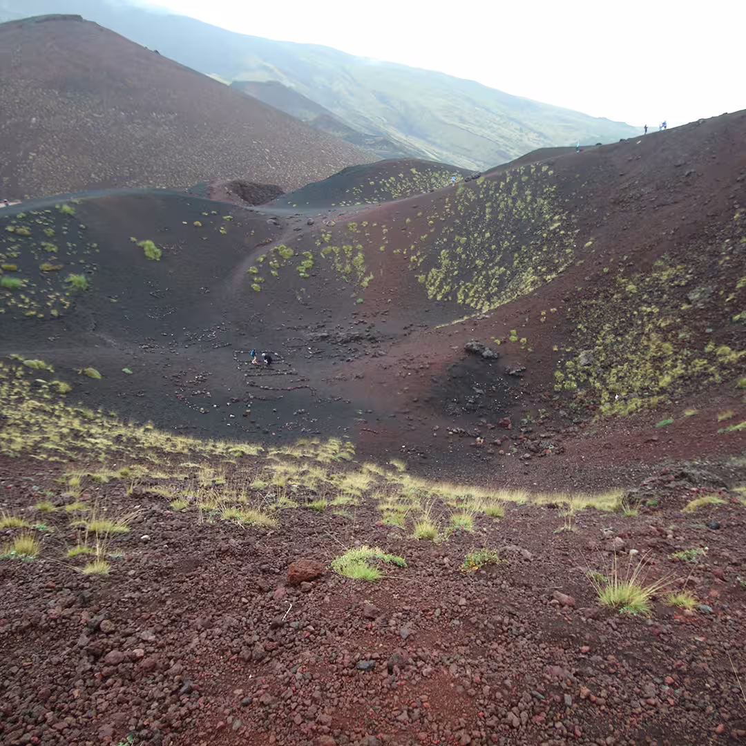 Volcanic landscape near Mount Etna, Sicily, Italy, during the Dyalog '15 event. The terrain consists of reddish-brown volcanic rock and black ash, with sparse tufts of green grass scattered across the slopes. In the distance, a few people are walking along the crater ridge. The background shows the slopes of Mount Etna, partially covered in mist or low clouds.