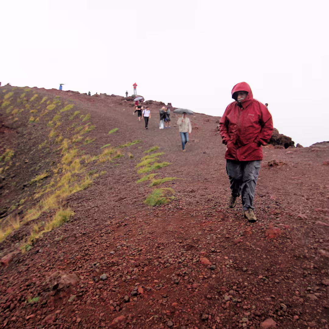 Geoff Streeter hiking near Mount Etna during the Dyalog '15 event in Sicily, Italy. He is wearing a red rain jacket with the hood up and grey trousers, walking along a rocky volcanic path. Behind him, several other delegates, some carrying umbrellas, are making their way along the trail. The ground is covered in reddish-brown volcanic rock with patches of green grass, and the sky is overcast.