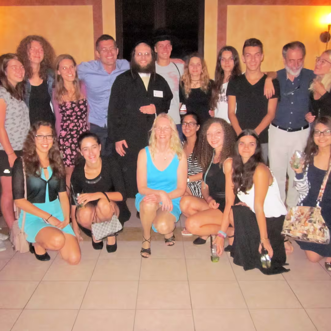 Group photo taken during the Dyalog '15 event in Sicily, Italy, showing a diverse group of delegates posing together indoors. The group includes men and women dressed in smart casual and semi-formal attire, with some holding drinks. Adám Brudzewsky, wearing traditional Orthodox Jewish attire, stands near the center. A woman in a bright blue dress is kneeling in the front row, smiling at the camera. The background features warm-toned walls with soft lighting.