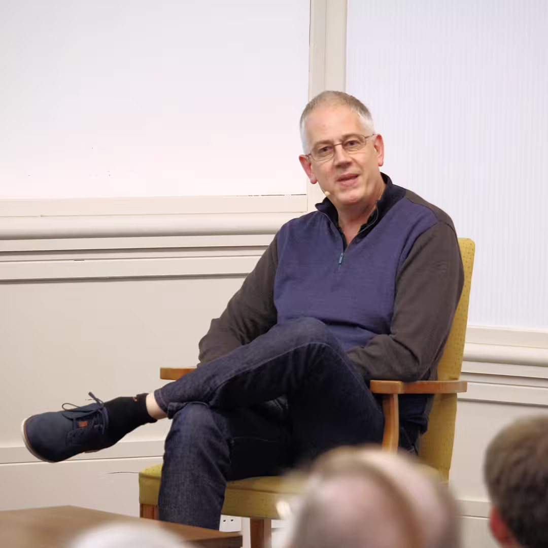 Andy Shires seated during Dyalog '16 in Glasgow, Scotland. He is wearing glasses, a navy blue and black pullover, dark jeans, and navy shoes with orange laces. He sits with his legs crossed on a yellow cushioned wooden chair, speaking while looking toward the audience. The background features a white wall and part of the conference room's paneling.