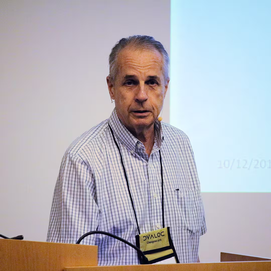 Charles Brenner presenting at Dyalog '16 in Glasgow, Scotland. He stands behind a wooden podium, wearing a checkered button-up shirt and a headset microphone. A Dyalog 2016 conference badge hangs from a lanyard around his neck. The background features a projected presentation slide with a partially visible date in the lower right corner.