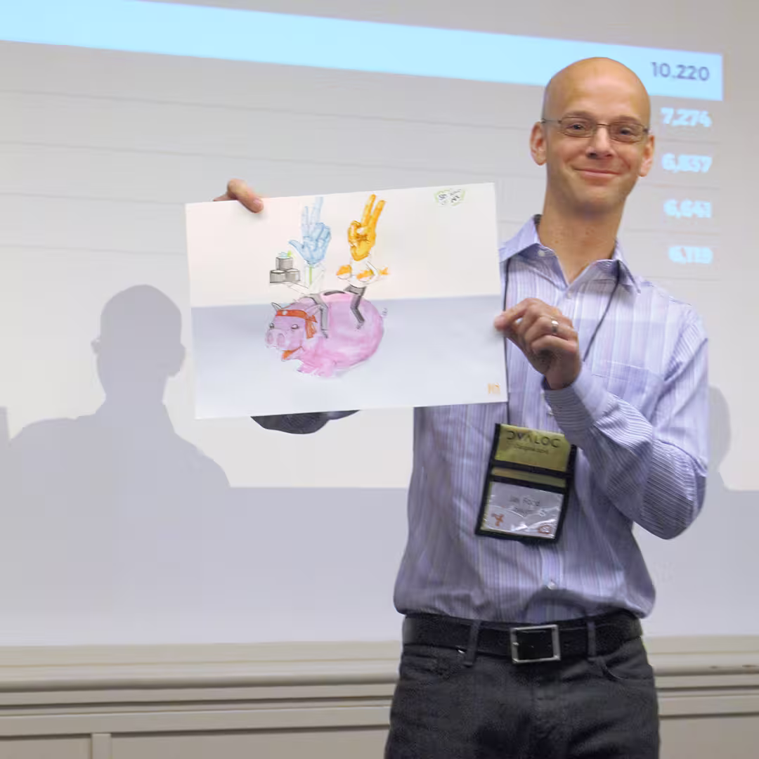Jay Foad presenting at Dyalog '16 in Glasgow, Scotland. He is holding up a colorful, hand-drawn illustration featuring a pink piggy bank with objects stacked on its back, including a peace sign, a robotic hand, and a small box. He is wearing a light purple striped shirt, dark trousers, and a conference badge. A presentation slide with numerical data is projected onto the screen behind him, and his shadow is visible on the wall.