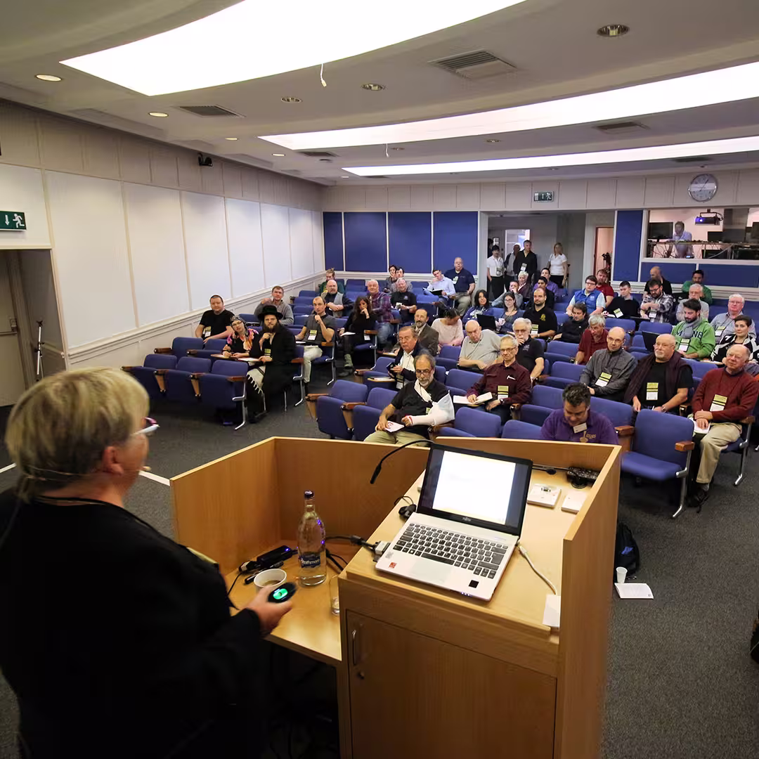 Gitte Christensen presenting at Dyalog '16 in Glasgow, Scotland. She stands at a wooden podium, holding a presentation remote and facing an audience seated in rows of purple chairs. A laptop and water bottle are on the podium. The audience consists of delegates, some taking notes, while others listen attentively. The conference room has white walls, blue panels, and a clock on the back wall. Some people are standing near the entrance, and a few are visible through a window into an adjacent room.