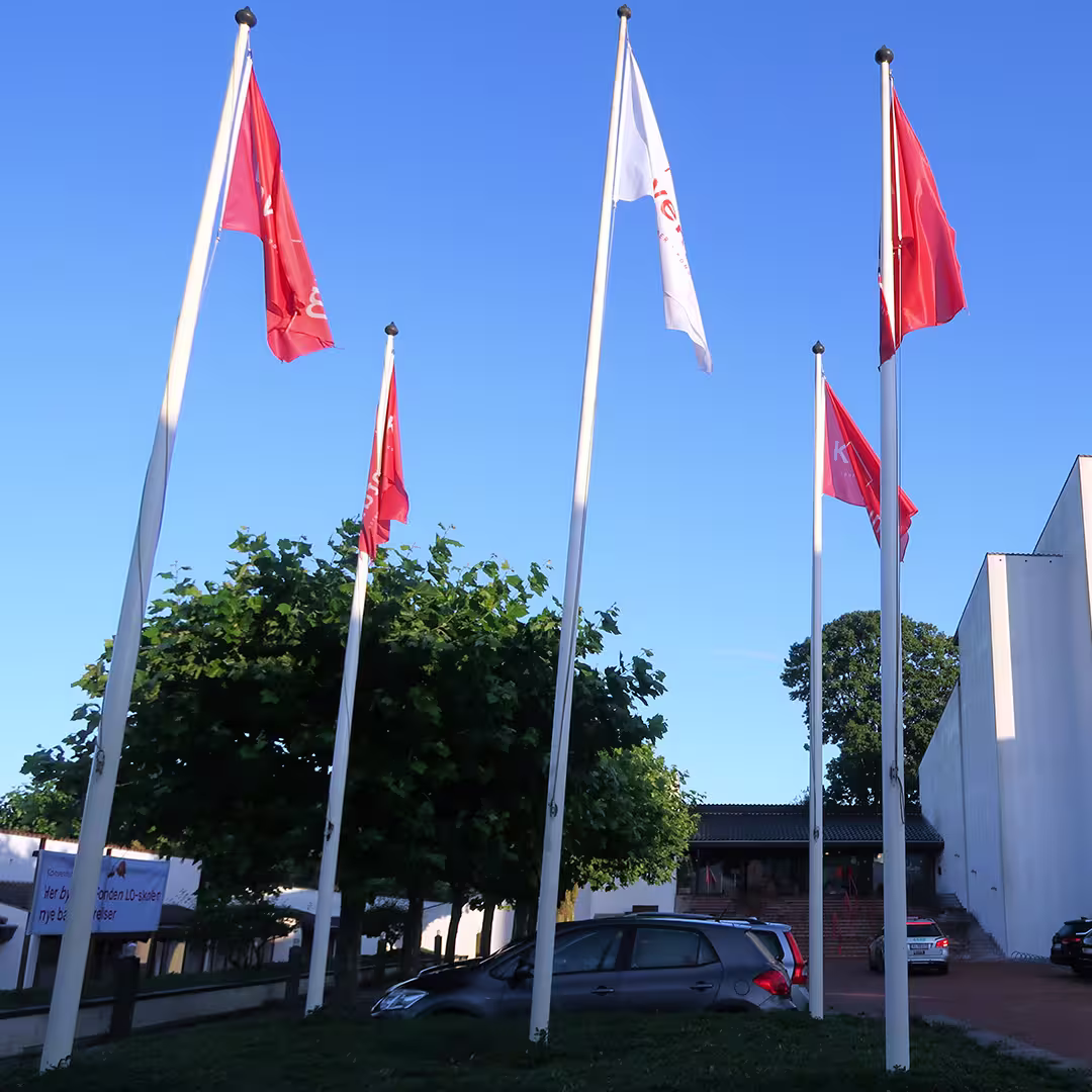 Five flagpoles stand in front of the Konventum Convention Centre in Elsinore, Denmark, during the Dyalog '17 event. Four red flags and one white flag, all bearing the Konventum logo, flutter in the breeze against a clear blue sky. Behind the flags, green trees partially obscure the white building facade, while two parked cars are visible in the foreground.