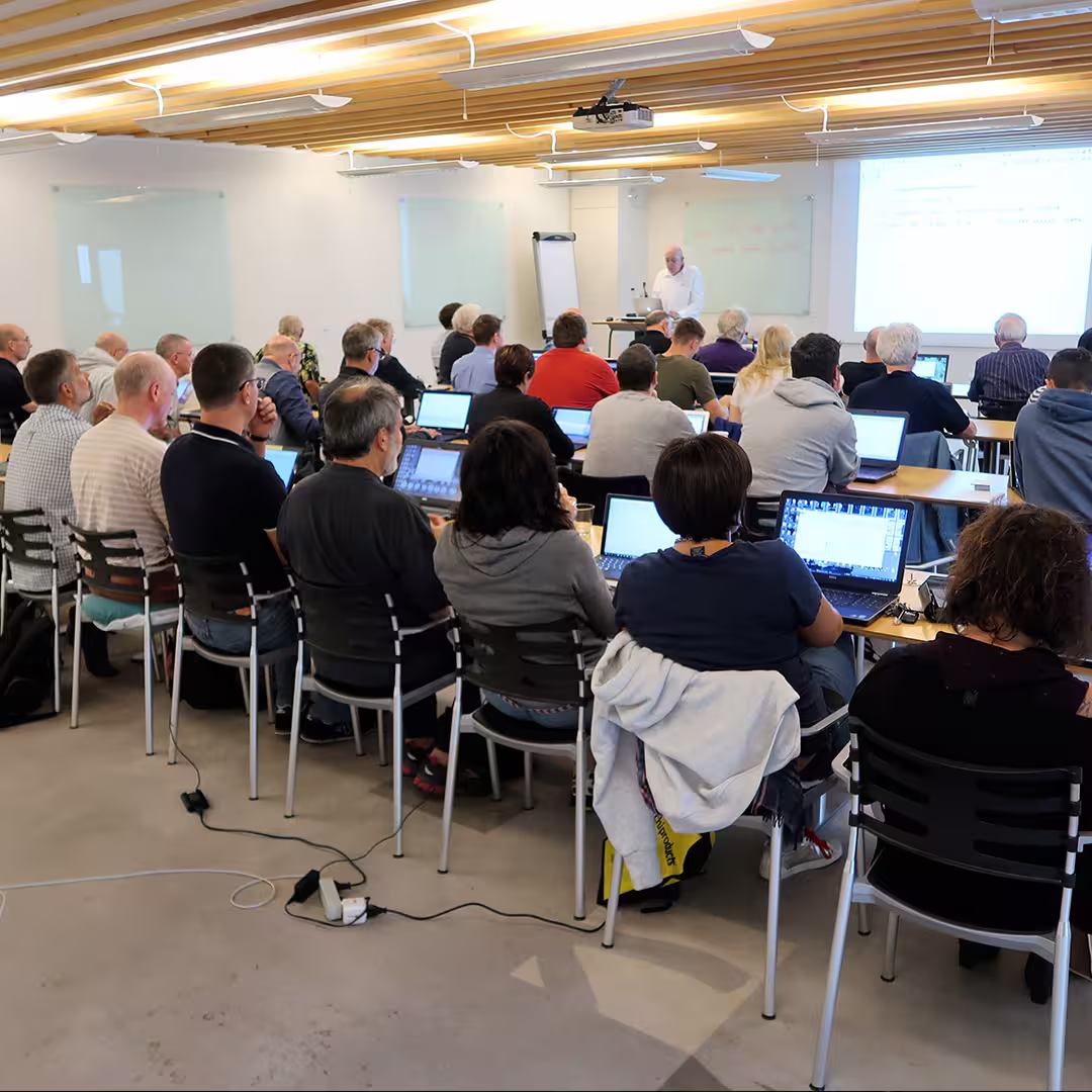 John Scholes runs a workshop at the Dyalog '17 event in Elsinore, Denmark. He stands at the front of a modern conference room, speaking to a full audience of delegates seated at long tables with laptops open. The room has a wooden slatted ceiling, white walls, and large glass panels used as whiteboards. A projector displays APL code on the screen behind him. Power cables run along the floor, connecting devices throughout the room.