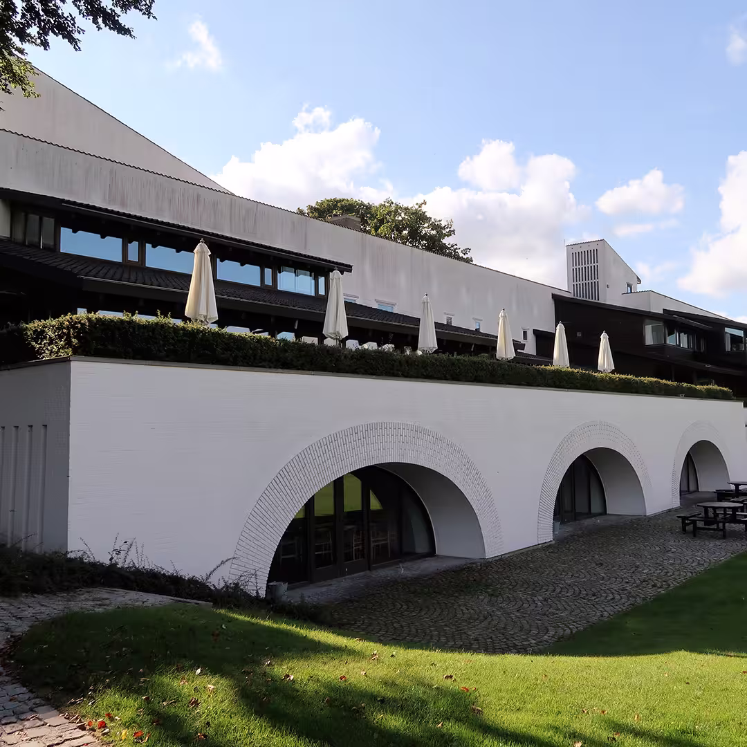 The exterior of the Konventum Convention Centre in Elsinore, Denmark, during the Dyalog '17 event. The modern white building features a row of large arched windows on the ground floor, with a cobblestone patio area in front. Above the arches, a terrace lined with green hedges and closed white patio umbrellas runs along the length of the building. The upper level has large windows and dark wooden cladding. The scene is framed by a well-maintained lawn, with trees and a partly cloudy sky in the background.