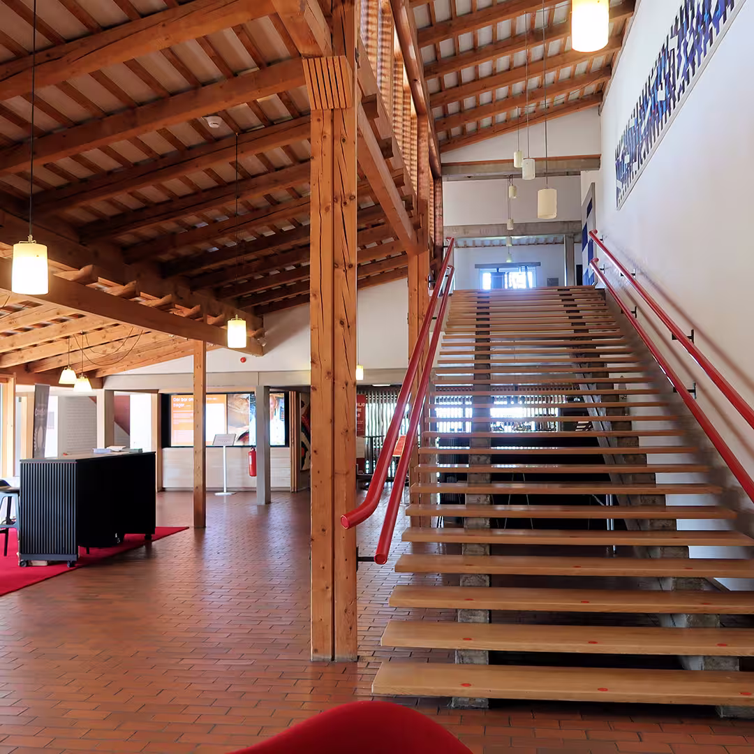 The interior of the Konventum Convention Centre in Elsinore, Denmark, during the Dyalog '17 event. The space features a large open area with exposed wooden beams, terracotta floor tiles, and hanging pendant lights. A wide wooden staircase with red handrails leads to the upper floor. To the left, the event registration desk sits on a red carpet. In the background, large windows allow natural light to stream in, while informational displays and artwork adorn the walls.