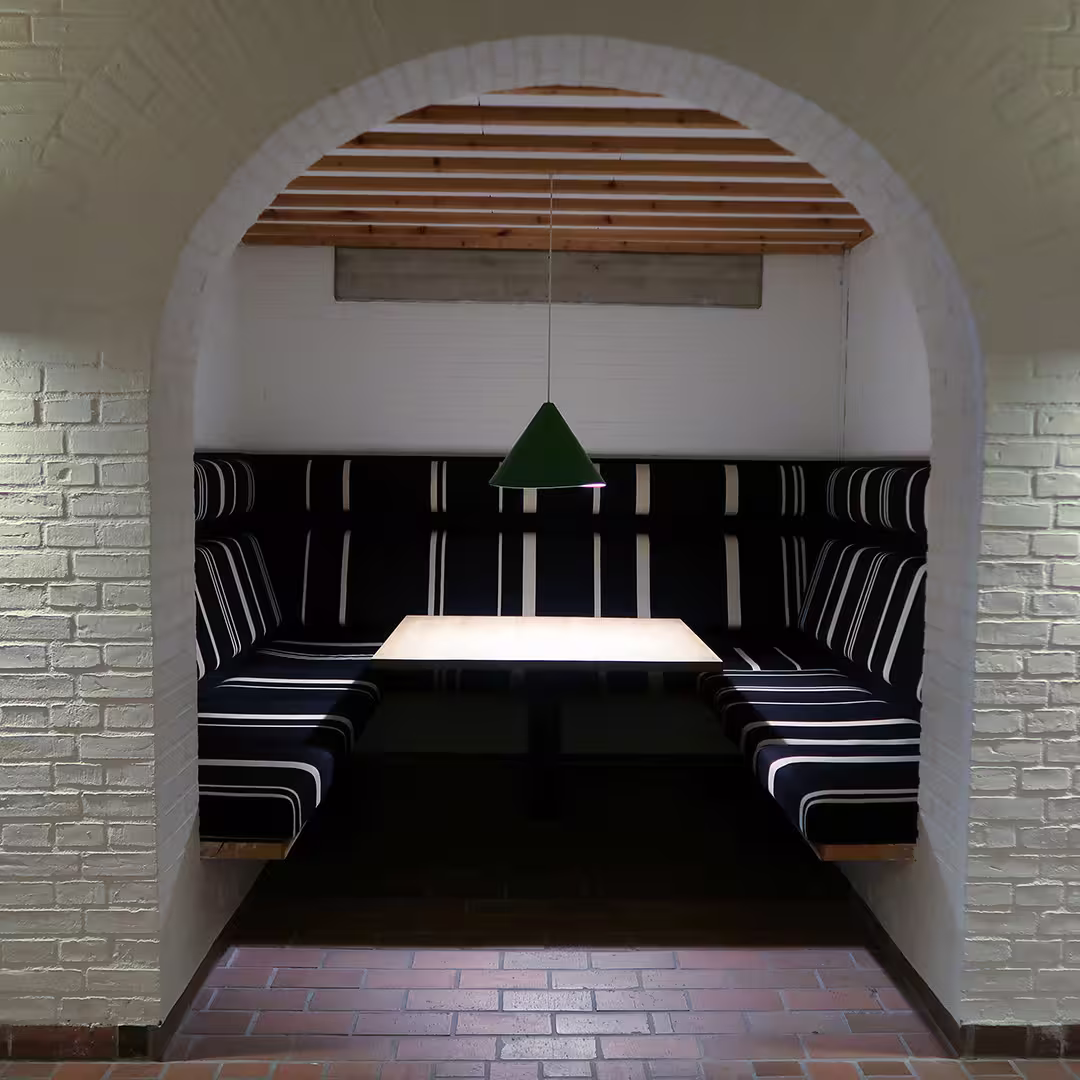 A cozy seating nook at the Konventum Convention Centre in Elsinore, Denmark, during the Dyalog '17 event. The nook is framed by a white brick archway and features built-in black benches with white vertical stripes, arranged in a U-shape around a light wooden table. A green pendant lamp hangs from the ceiling, casting soft light onto the table. The floor is made of reddish-brown tiles, and the back wall is white with exposed wooden ceiling beams.