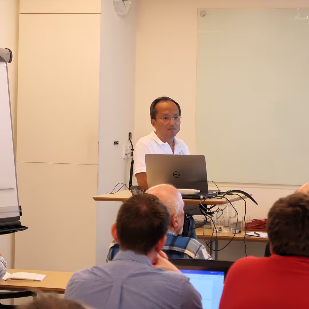 Roger Hui running a workshop at the Dyalog '17 event in Elsinore, Denmark. He stands behind a lectern with a Dell laptop, wearing a white Dyalog-branded polo shirt and speaking to an audience of delegates seated in rows. Some attendees are using laptops while others take notes. A flip chart stands to the side, and a whiteboard is mounted on the wall behind Roger. Cables and electronic equipment are arranged on the desk.