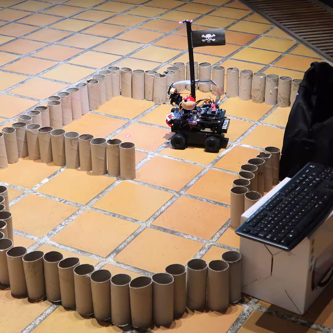 A Raspberry Pi-powered robot decorated as a pirate ship moves within a maze made of upright cardboard tubes at the Dyalog '17 event in Elsinore, Denmark. The robot features a black flag with a skull and crossbones, a pirate figurine, and various electronic components mounted on its chassis. A red laser pointer shines on the floor in front of the robot, guiding its path. A keyboard rests on a white box nearby, likely for controlling the robot. The setup is placed on a tiled floor in an indoor conference space.