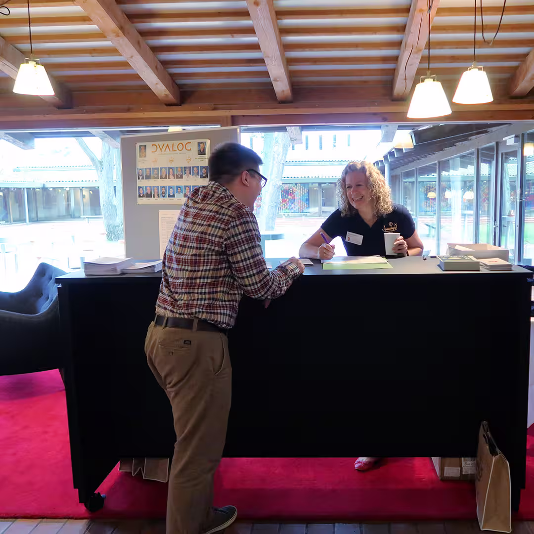 Karen Shaw of Dyalog Ltd stands behind the registration desk at the Dyalog '17 event in Elsinore, Denmark, smiling while assisting a delegate wearing a plaid shirt and glasses. She holds a coffee cup in one hand and a pen in the other, ready to check in the attendee. The desk is covered with event materials, and a Dyalog staff photo board is displayed behind them. Large windows reveal an outdoor courtyard with trees and modern architecture. The scene is lit by hanging lights under a wooden ceiling, and a red carpet runs beneath the desk.