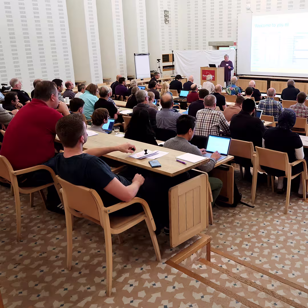 Gitte Christensen presents at the Dyalog '17 event in Elsinore, Denmark, standing behind a Konventum-branded podium. She is speaking to a full audience seated in tiered wooden rows, with many delegates taking notes or using laptops. The presentation slide on the screen reads "Welcome to you all." The room features light-colored walls, patterned carpet, and a white backdrop behind the speaker. A flip chart and video camera stand to the side of the room.