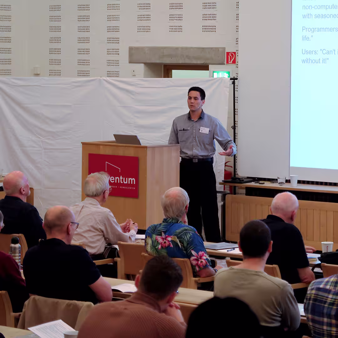 Aaron Hsu presents at the Dyalog '17 event in Elsinore, Denmark, standing behind a Konventum-branded podium. He is wearing a light grey shirt and black trousers, gesturing with his right hand while speaking. Behind him, a white backdrop covers part of the wall. In the foreground, several delegates are seated, some taking notes, including a man in a colourful Hawaiian shirt. The presentation slide on the right side of the image features user quotes, including "Can't live without it!"