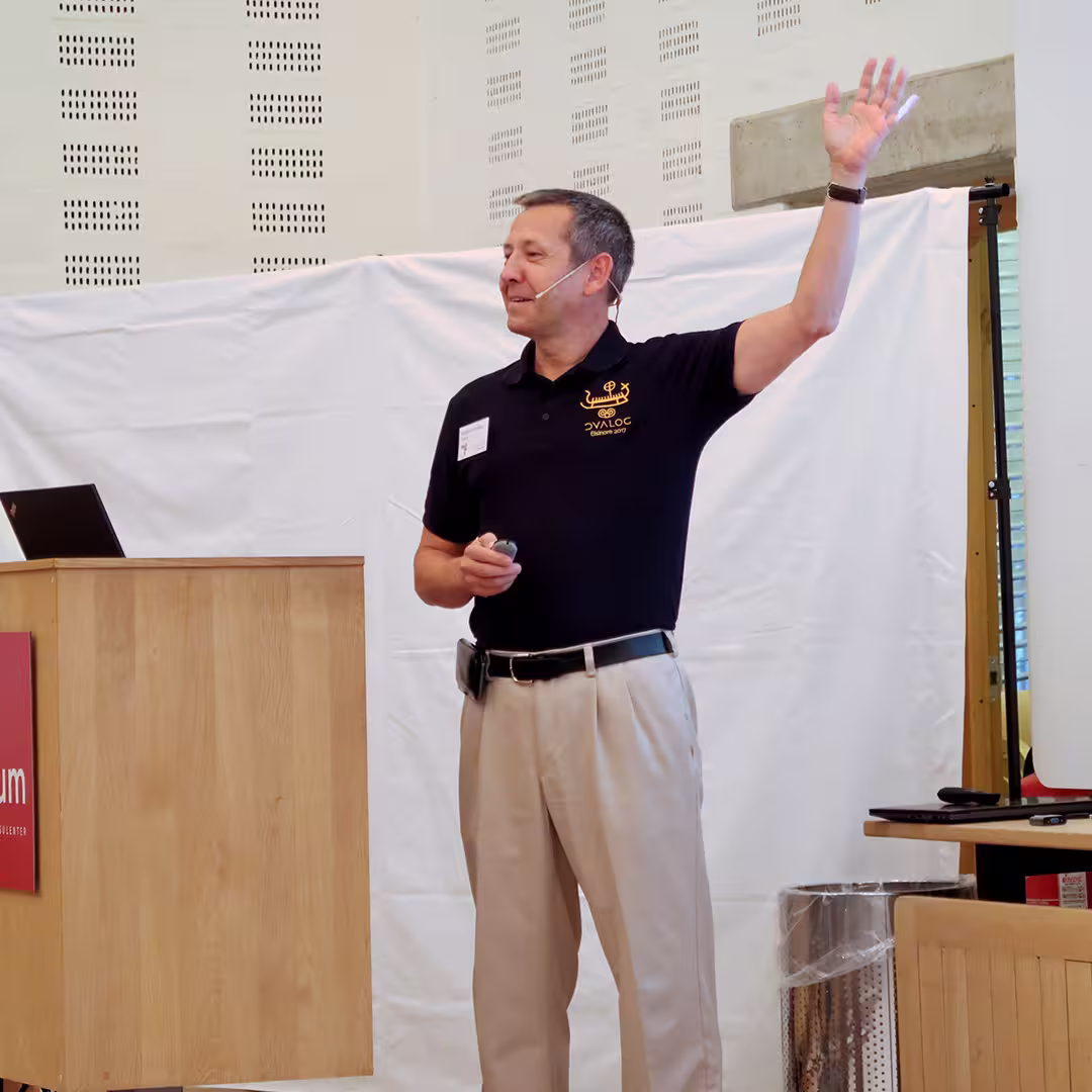 Morten Kromberg presents at the Dyalog '17 event in Elsinore, Denmark, standing beside a Konventum-branded podium. He is wearing a black Dyalog polo shirt with an embroidered logo and beige trousers. Speaking into a headset microphone, he raises his right hand while holding a presentation remote in his left. A white backdrop hangs behind him, and a stainless steel bin is visible near the stage.