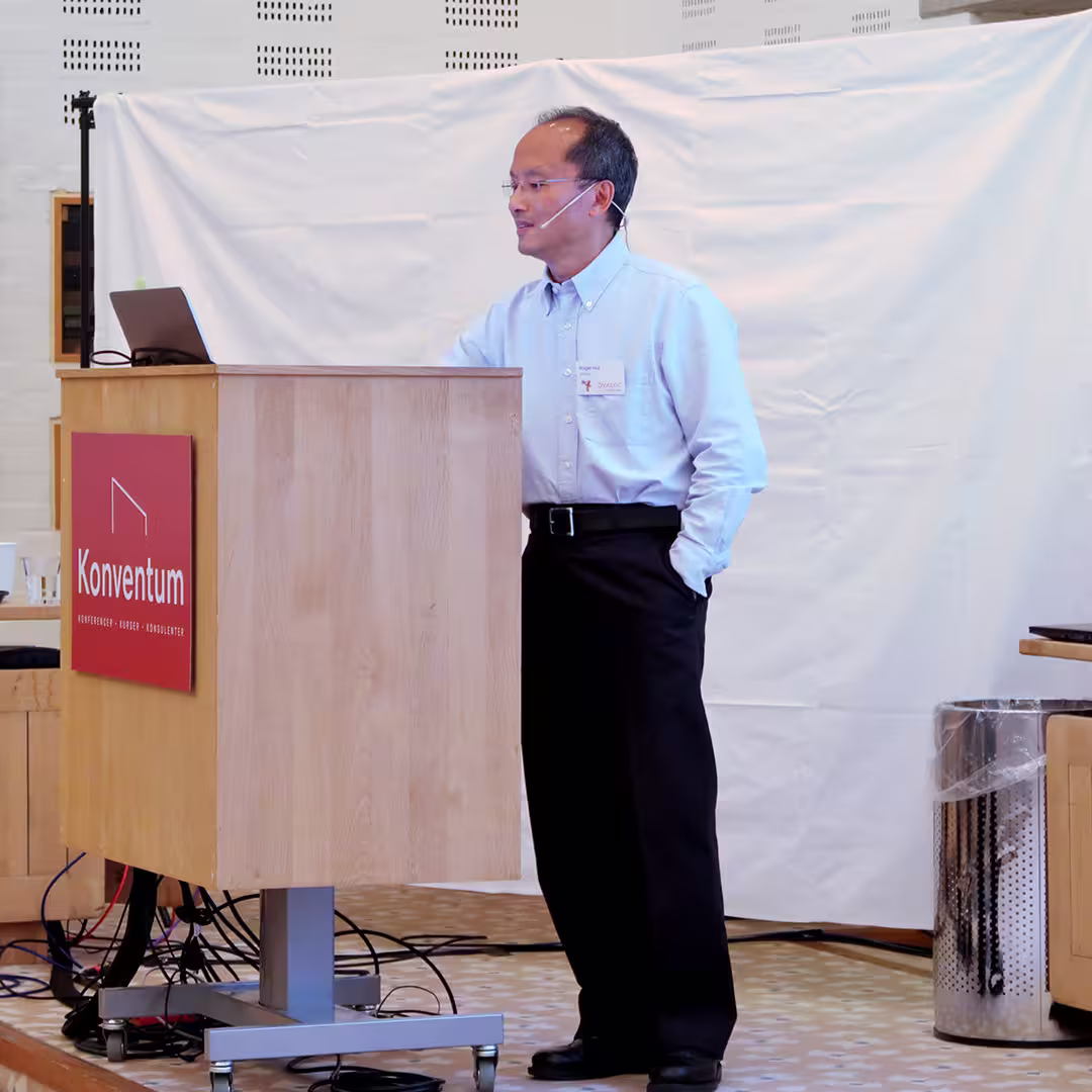 Roger Hui stands behind a wooden podium branded with the Konventum logo, delivering a presentation at the Dyalog '17 event in Elsinore, Denmark. He is wearing a light blue shirt and black trousers, speaking into a headset microphone while looking at his laptop. A white backdrop hangs behind him, and a stainless steel bin is visible to the side of the stage. Cables run across the floor, connecting the podium to the presentation equipment.