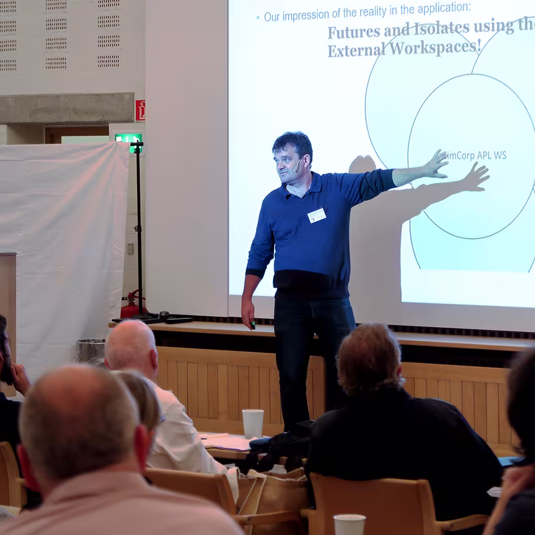 A presenter at the Dyalog '17 event in Elsinore, Denmark, stands in front of a large projection screen, gesturing toward a Venn diagram on the slide. The slide title reads, "Futures and Isolates using External Workspaces!" The presenter, wearing a dark blue sweater and jeans, has a conference badge clipped to his shirt. Audience members are seated in the foreground, some leaning forward attentively. The room features light-colored walls and wooden furnishings typical of the Konventum Convention Centre.
