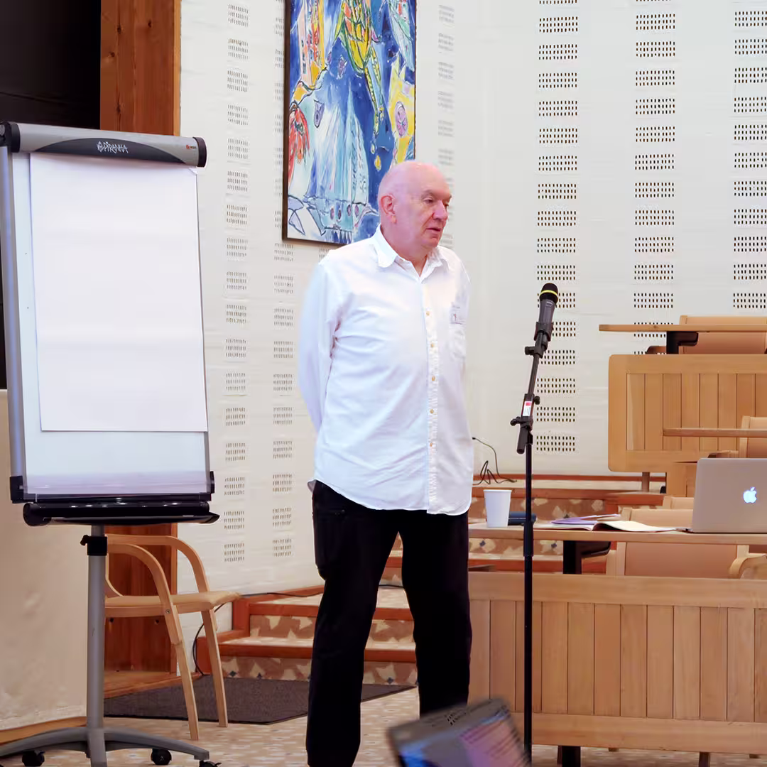 John Scholes stands on stage during the Dyalog '17 event at the Konventum Convention Centre in Elsinore, Denmark. He is wearing a white shirt and dark trousers, speaking into a microphone on a stand. To his left, a blank flip chart stands on a wheeled easel. Behind him, the light-colored walls feature a colorful abstract painting. A table with a laptop, papers, and a coffee cup is visible in the background, along with the room's tiered wooden seating.