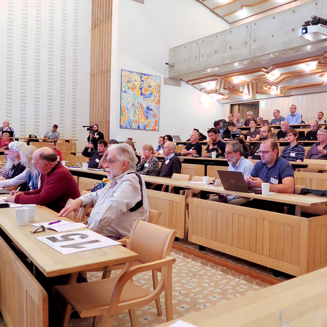 Delegates at the Dyalog '17 event in Elsinore, Denmark, sit in tiered wooden seating inside the Konventum Convention Centre, attentively listening to a presentation. Some take notes, while others use laptops or hold cups of coffee. A man in the foreground, wearing glasses and a light shirt, sits at a desk with a sign reading "15" in front of him. The room features modern architecture with wooden ceiling panels and light walls. A colorful abstract painting hangs on the wall, and a videographer operates a camera near the back of the room.