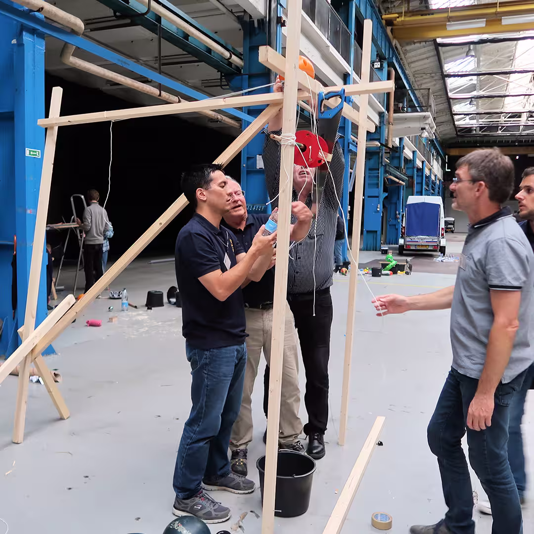A group of delegates at the Dyalog '17 event in Elsinore, Denmark, collaborate on constructing a wooden contraption inside an industrial-style venue with high ceilings and blue metal beams. Four people are actively working together, adjusting strings and securing components. One person holds a plastic bottle while another reaches up to adjust a pulley system. A black bucket sits on the floor nearby, surrounded by scattered tools and materials. A van is visible in the background near the open end of the venue.